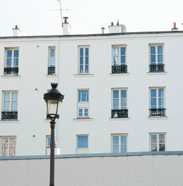 Nathalie F. · 07/02/2026 · 2981 x 3033 Cette photographie met en scène un immeuble parisien typique de style haussmannien. Le sujet principal est l’architecture, avec ses fenêtres symétriques et balcons en fer forgé. La composition est équilibrée, centrée sur un réverbère ancien en premier plan.Le cadrage est serré, mettant en valeur les détails architecturaux. La lumière est douce et naturelle, légèrement voilée, créant une ambiance sobre. Les couleurs dominantes sont les tons pastel (bleu pâle, blanc) et le gris clair, renforçant une atmosphère calme et intemporelle. L’ensemble dégage une élégance classique et une touche nostalgique.