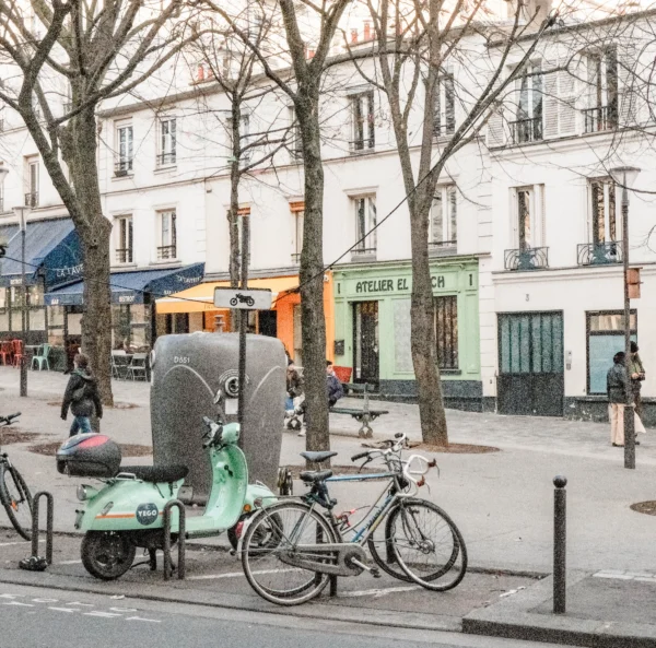 Nathalie F. · 07/02/2026 · 3895 x 3847 Cette photographie met en scène une rue urbaine animée. Le **sujet principal** est un vélo-cargo vert clair garé près d’un arbre, avec une caisse de déménagement dessus.La **composition** est équilibrée, avec des éléments architecturaux (bâtiments, enseignes) et des arbres dénudés en arrière-plan. Le **cadrage** est large, incluant une partie du trottoir et de la chaussée.La **lumière** est naturelle et douce, typique d’un jour hivernal ou automnal. Les **couleurs dominantes** sont les tons neutres (gris, beige) adoucis par les touches de vert et bleu.Le **style** est réaliste et documentaire, capturant une scène de vie quotidienne. L’**ambiance** est calme et urbaine, évoquant une ville européenne.