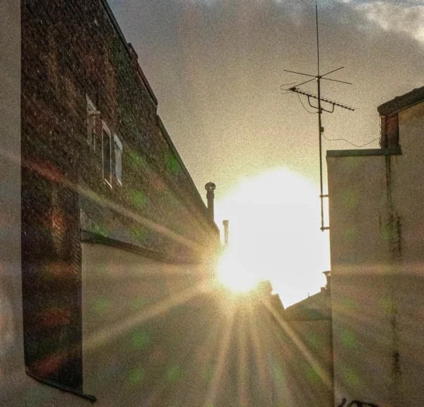 Nathalie F. · 07/02/2026 · 3234 x 3098 Cette photographie capture un coucher de soleil dans une ruelle urbaine étroite. Le **sujet principal** est le soleil bas, créant des **rayons lumineux** qui traversent l’espace. La **composition** est symétrique, avec des bâtiments de chaque côté encadrant la lumière.Le **cadrage** est serré, mettant en valeur la perspective et les reflets. La **lumière** est douce mais intense, avec des contrastes marqués. Les **couleurs dominantes** sont des tons chauds (oranges, jaunes) et des teintes grises-brunâtres des murs.Le **style** est réaliste et légèrement flou, évoquant une ambiance **nostalgique et paisible**, presque onirique.