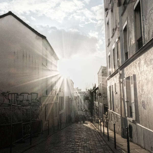 Nathalie F. · 07/02/2026 · 4160 x 4160 Cette photographie capture une **rue étroite et pavée** en ville, avec des **immeubles anciens** de part et d'autre. Le **sujet principal** est la lumière du soleil filtrant entre les bâtiments, créant un jeu de rayons lumineux.La **composition** est symétrique, avec une perspective convergente vers l’horizon. Le **cadrage** est serré, mettant en valeur les détails architecturaux et les graffitis. La **lumière** est douce et contrastée, avec des ombres marquées.Les **couleurs dominantes** sont des tons gris-beige, adoucis par les touches de lumière dorée. Le style est **réaliste et légèrement mélancolique**, évoquant une **ambiance calme et nostalgique**.