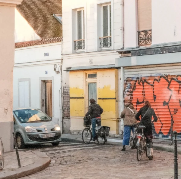 Nathalie F. · 07/02/2026 · 3369 x 3341 Cette photographie capture une scène urbaine animée. Le sujet principal est un groupe de cyclistes discutant près d’un bâtiment tagué. La composition met en valeur une rue étroite avec des bâtiments en fond, dont un mur jaune et un autre couvert de graffitis orange et noirs.Le cadrage est serré, centré sur les cyclistes et les détails architecturaux. La lumière semble naturelle, douce et légèrement tamisée, typique d’un jour couvert ou matinal. Les couleurs dominantes sont les tons neutres (gris, beige) contrastant avec les jaunes et oranges vifs des murs.L’ambiance est réaliste et quotidienne, reflétant une vie urbaine authentique et décontractée.