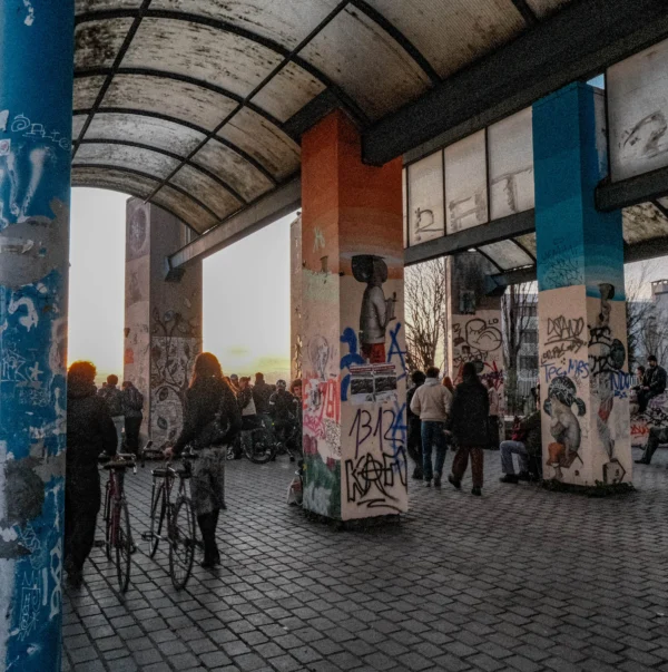 Nathalie F. · 07/02/2026 · 4032 x 4050 Cette photographie montre un espace urbain couvert, probablement une gare ou un passage piéton, décoré de graffitis colorés. Le sujet principal est l’interaction entre les personnes et cet environnement artistique.La composition met en valeur les colonnes et les murs tagués, encadrant une scène animée. Le cadrage inclut des détails architecturaux et humains, créant une ambiance dynamique. La lumière, tamisée et naturelle, provient probablement de l’extérieur, adoucissant les contrastes.Les couleurs dominantes sont vives (bleu, orange, vert) grâce aux graffitis, contrastant avec les tons neutres des structures. Le style est urbain et street art, évoquant une ambiance jeune, créative et légèrement underground.