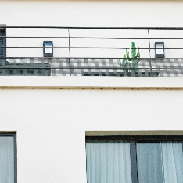 Nathalie F. · 21/02/2026 · 4160 x 4160 Cette photographie met en scène un bâtiment moderne avec un **sujet principal** : une statue verte en forme de cactus ou de créature stylisée, posée sur un balcon.La **composition** est épurée, avec un **cadrage serré** sur la façade. Les **lignes horizontales** (balcon, fenêtres) et verticales (murs) structurent l'image. La **lumière** est douce et naturelle, créant des ombres légères.Les **couleurs dominantes** sont neutres (blanc, gris) avec une touche de vert vif pour la statue. Le **style** est minimaliste et contemporain, évoquant une **ambiance** urbaine et moderne, presque surréaliste grâce à l'élément végétal ou organique contrastant avec l'architecture.