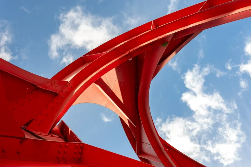Daniel · 04/02/2026 · 5293 x 3529 Cette photographie met en scène un **sujet architectural abstrait**, probablement une structure métallique rouge en forme d’arcs entrelacés. La **composition** joue sur les lignes dynamiques et les courbes, créant un mouvement visuel. Le **cadrage serré** isole les éléments, accentuant leur forme et leur texture.La **lumière** naturelle, probablement en plein jour, éclaire uniformément la structure, mettant en valeur ses contours nets. Les **couleurs dominantes** sont le rouge vif et le bleu du ciel, contrastant avec les nuages blancs. Le style est épuré et moderne, évoquant une **ambiance industrielle et artistique**, presque sculpturale.
