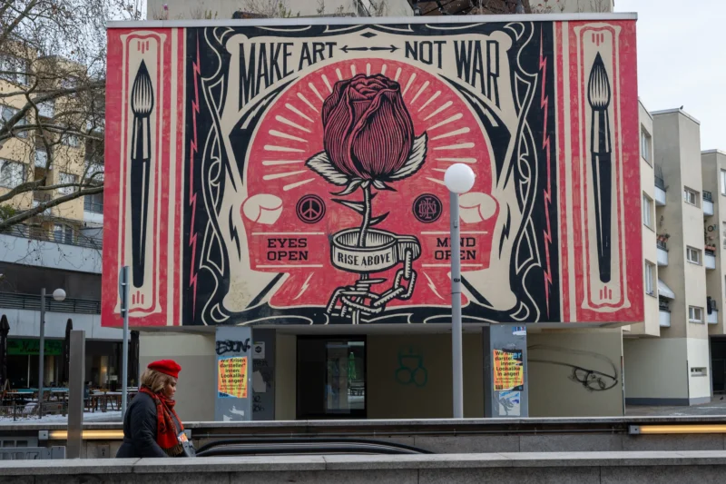 Daniel · 19/02/2026 · 5141 x 3427 Cette photographie montre un mural coloré sur un bâtiment, représentant un visage stylisé avec des éléments symboliques (couteaux, fleurs, yeux). Le texte "Make Art Not War" est visible en haut.La composition est verticale, avec un cadrage serré sur l’affiche. Les couleurs dominantes sont le rouge, le noir et le blanc, créant un contraste fort. La lumière semble naturelle, éclairant le panneau de manière uniforme.Le style est artistique et engagé, évoquant un message pacifiste. L’ambiance est à la fois puissante et provocante, renforcée par les éléments graphiques percutants.