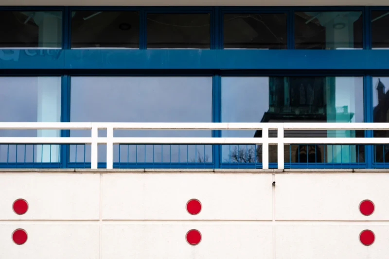 Daniel · 24/02/2026 · 5451 x 3634 Cette photographie montre principalement une partie d’un bâtiment moderne avec une façade blanche et des balustrades bleues. Le sujet secondaire est une série de petits cercles rouges alignés horizontalement.La composition est épurée, axée sur des lignes horizontales et géométriques. Le cadrage est serré, centré sur ces éléments architecturaux.Les couleurs dominantes sont le blanc, le bleu et le rouge vif. La lumière semble naturelle, mais diffuse, créant un contraste doux.Le style est minimaliste et contemporain, évoquant une ambiance sobre et moderne, presque industrielle.