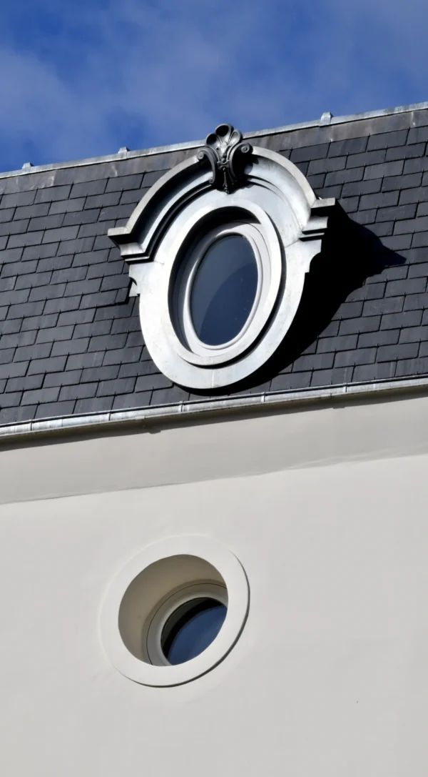 Vincent · 21/02/2026 · 2900 x 5253 Cette photographie met en scène un **détail architectural** : une **lucarne ovale** surmontée d’un petit acrotère orné, sur un toit d’ardoise.La **composition** est centrée sur cet élément, avec un **cadrage serré** qui isole la structure. La lumière, **naturelle et vive**, crée des contrastes nets entre ombres et zones éclairées. Les **couleurs dominantes** sont les tons gris-bleu des ardoises, le blanc crème du mur et les teintes claires de la lucarne.Le style est **minimaliste et épuré**, évoquant une architecture classique ou traditionnelle. L’ambiance est **calme et élégante**, renforcée par la netteté des formes et la clarté du ciel.