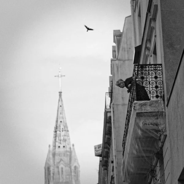 Cyrille · 2026 x 2026 Cette photographie en noir et blanc met en scène un homme penché sur un balcon, semblant observer ou travailler. Le sujet principal est cet individu, encadré par une architecture urbaine et une tour gothique en arrière-plan.La composition joue sur les lignes verticales des bâtiments et la perspective, créant une ambiance contemplative. Le cadrage serré isole le personnage tout en intégrant des éléments architecturaux suggestifs.La lumière, diffuse et tamisée, accentue les contrastes et donne une touche mélancolique. Les tons dominants, gris et noirs, renforcent une atmosphère sobre et introspective, typique d’un style photographique documentaire ou artistique.