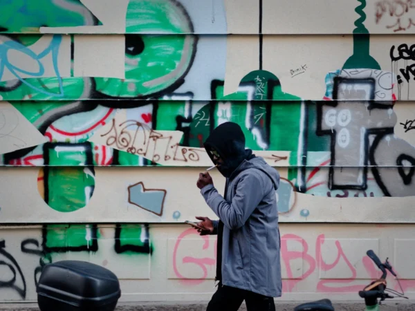 Cyrille · 2026 x 1520 Cette photographie met en scène un individu vêtu d’une veste longue et d’un bonnet, utilisant un téléphone portable devant un mur tagué. Le sujet principal est légèrement flou, attirant l’attention sur les graffitis colorés derrière lui.La composition joue sur le contraste entre le personnage et le mur vibrant. Le cadrage est serré, centré sur le sujet et le graffiti. La lumière est naturelle, douce mais contrastée, mettant en valeur les couleurs vives du mur.Les couleurs dominantes sont vertes, rouges et noires, typiques des graffitis urbains. Le style est réaliste et documentaire, capturant une scène de rue authentique. L’ambiance est à la fois dynamique et mélancolique, reflétant une vie urbaine.