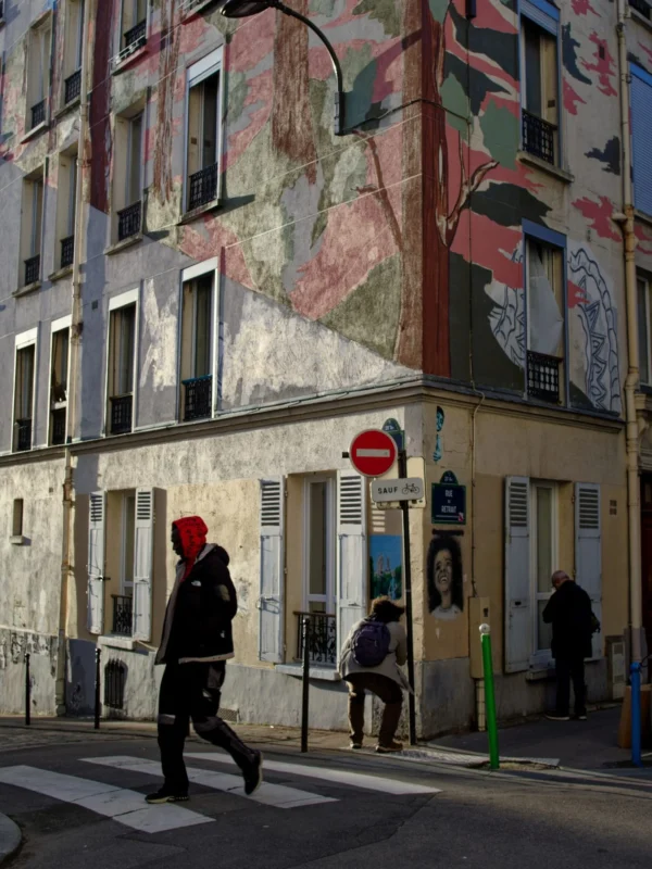 Cyrille · 1520 x 2026 Cette photographie capture une scène urbaine animée. Le **sujet principal** semble être un immeuble aux façades colorées et dégradées, couvertes de fresques murales vieillissantes. La **composition** met en valeur un angle de rue avec des passants, dont une personne en rouge marchant vers l’appareil.Le **cadrage** est serré, centré sur l’intersection et les bâtiments. La **lumière** est naturelle, avec des ombres marquées, suggérant un ensoleillement matinal ou tardif. Les **couleurs dominantes** sont des tons pastel (bleu, rose) contrastant avec le gris et le vert des éléments urbains.Le **style** est documentaire et réaliste, évoquant une ambiance **urbaine authentique**, entre vie quotidienne et traces artistiques éphémères.