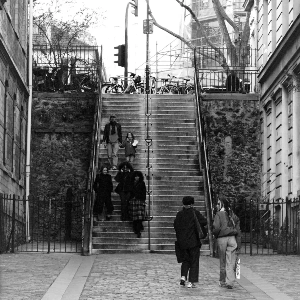 Cyrille · 2026 x 2026 Cette photographie en noir et blanc capture une montée d'escaliers urbaine. Le sujet principal est un groupe de personnes, dont deux en premier plan qui marchent vers les escaliers. La composition met en valeur les escaliers centraux, encadrés par des bâtiments et des arbres.Le cadrage est serré, guidant le regard vers le haut. La lumière est naturelle, contrastant les ombres et les formes. Les couleurs dominantes, bien que monochromes, jouent sur les nuances de gris et de noir.Le style est documentaire et réaliste, évoquant une ambiance quotidienne et urbaine, presque mélancolique.