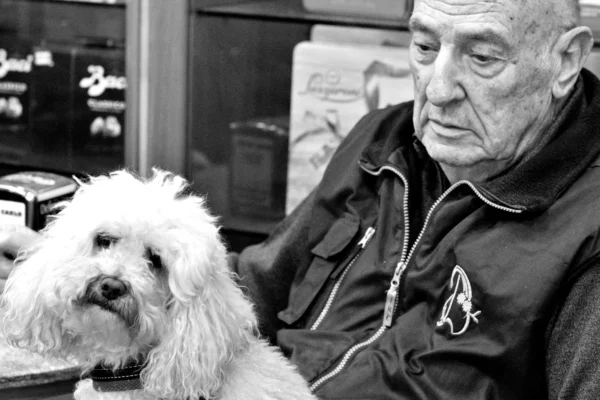 Fabienne · 13/02/2026 · 4047 x 2698 Cette photographie met en scène un homme âgé comme sujet principal, assis et accompagné d'un petit chien blanc. La composition est simple et intime, avec un cadrage serré sur les deux personnages. L'arrière-plan, légèrement flou, montre des étagères remplies de livres et d'objets.La lumière est douce et naturelle, créant des contrastes modérés. En noir et blanc, les tons dominants varient du clair au foncé, accentuant les textures et les expressions. Le style est documentaire et réaliste, évoquant une ambiance chaleureuse et nostalgique.