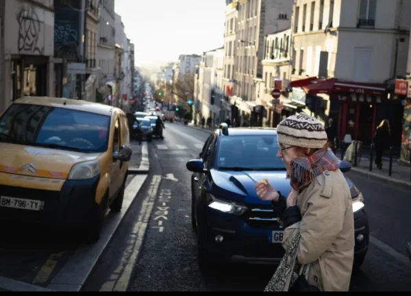 Muriel · 1080 x 777 Cette photographie montre une personne vêtue d’un manteau clair et d’un bonnet à pompon, tenant un sac, comme sujet principal. Elle se tient sur un trottoir urbain, en retrait d’un trafic dense.La composition est dynamique, avec une rue étroite bordée de bâtiments et de voitures. Le cadrage est serré sur la personne, mais inclut aussi l’arrière-plan animé. La lumière est naturelle, légèrement tamisée, créant des ombres douces. Les couleurs dominantes sont les tons neutres (gris, beige) avec des éclats de couleurs vives (voitures jaunes, rouge du feu).Le style est documentaire, capturant une scène de vie quotidienne. L’ambiance est urbaine et réaliste.