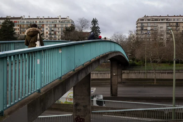 Muriel · 2048 x 1365 Cette photographie met en scène un **sujet principal** : un homme en imperméable marchant sur un **pont métallique vert**. La **composition** est simple, avec une perspective légèrement basse, mettant en valeur le pont et l’architecture urbaine en arrière-plan.Le **cadrage** est serré sur le sujet, tout en intégrant l’environnement. La **lumière** est naturelle mais tamisée, évoquant un jour gris et froid. Les **couleurs dominantes** sont les tons verts du pont, gris des bâtiments et du ciel, avec des touches de brun et de noir.Le **style** est réaliste et documentaire. L’**ambiance** est urbaine, mélancolique et calme, renforcée par le ciel couvert et l’absence de mouvement.