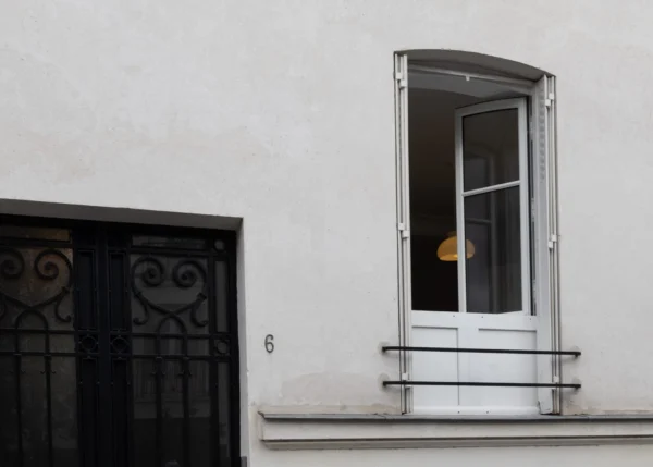Muriel · 2048 x 1463 Cette photographie met en scène une fenêtre ouverte sur un mur blanc, encadrée par un petit balcon en fer forgé. Le sujet principal est la lumière dorée traversant la fenêtre, éclairant une partie de l’intérieur.La composition est épurée, centrée sur la fenêtre et son cadre architectural. Le cadrage est serré, isolant ces éléments. La lumière naturelle, douce et chaude, crée un contraste avec l’ombre environnante. Les couleurs dominantes sont le blanc du mur, le gris du métal et les tons chauds de la lumière.Le style est minimaliste et photographique, évoquant une ambiance calme et introspective.
