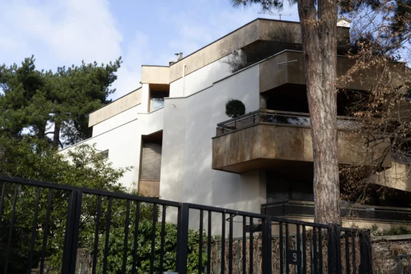 Muriel · 2048 x 1365 Cette photographie montre un immeuble moderne de deux étages, sujet principal. La composition met en valeur sa façade sobre et ses balcons en saillie. Le cadrage, légèrement bas, est encadré par une clôture métallique en premier plan.La lumière naturelle, douce, semble matinale ou tardive, créant des ombres légères. Les couleurs dominantes sont les tons beiges et blancs de l’immeuble, contrastés avec le vert des arbres et la clôture noire. Le style architectural est épuré et contemporain. L’ambiance est calme et résidentielle.