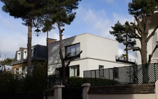 Muriel · 2048 x 1280 Cette photographie met en scène une maison moderne aux lignes épurées, sujet principal de l'image. La composition montre une façade claire avec des balcons en bois et des murs blancs, entourée de végétation (arbres et buissons). Le cadrage inclut partiellement des maisons voisines et un lampadaire.La lumière est naturelle, douce, avec des ombres légères, suggérant un jour nuageux. Les couleurs dominantes sont les tons blancs, gris et verts, renforçant une ambiance calme et contemporaine. Le style architectural est minimaliste et élégant, évoquant une ambiance résidentielle paisible et raffinée.