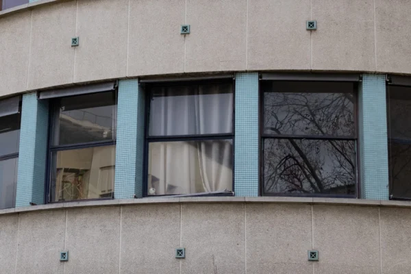 Muriel · 2048 x 1365 Cette photographie montre une partie d’un bâtiment avec trois fenêtres alignées sous un auvent arrondi. Le sujet principal est l’architecture sobre et les fenêtres aux cadres teintés en bleu-vert.La composition est symétrique, avec un cadrage serré sur les ouvertures. La lumière naturelle éclaire les surfaces, créant des contrastes doux. Les couleurs dominantes sont les tons gris-beige du mur, les teintes bleutées des cadres et les nuances claires des rideaux.Le style est épuré, typique d’une architecture moderne ou fonctionnaliste. L’ambiance est calme et minimaliste, renforcée par les arbres dénudés reflétés dans les vitres.