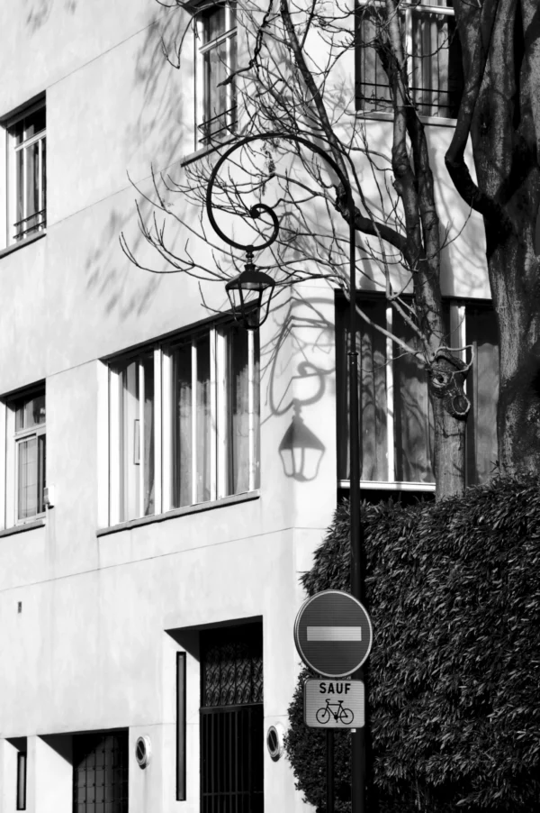 Thomas · 21/02/2026 · 2048 x 3080 Cette photographie en noir et blanc met en scène une façade d'immeuble urbain classique. Le sujet principal est un bâtiment avec des fenêtres alignées et une balustrade en fer forgé. La composition inclut un lampadaire et un panneau de signalisation "sens interdit" en bas à droite.Le cadrage est serré, centré sur l’architecture et les détails architecturaux. La lumière semble naturelle, avec des contrastes marqués créant des ombres nettes. Les couleurs dominantes sont les tons de gris, typiques du noir et blanc, renforçant une ambiance sobre et intemporelle. Le style est documentaire et esthétique, évoquant une atmosphère calme et légèrement mélancolique.