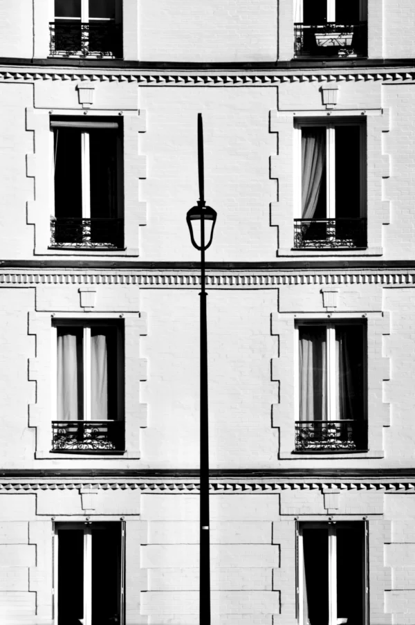 Thomas · 21/02/2026 · 2048 x 3081 Cette photographie met en scène une façade d’immeuble classique. Le sujet principal est un lampadaire central encadré par des fenêtres symétriques, disposées en grille régulière.Le cadrage est serré, centré sur ces éléments architecturaux. La lumière, naturelle et contrastée, accentue les ombres et les lignes géométriques. En noir et blanc, les tons dominants varient du blanc au gris foncé, renforçant le côté minimaliste et intemporel.Le style est épuré, presque graphique, évoquant une ambiance sobre et élégante, typique de l’architecture urbaine européenne.