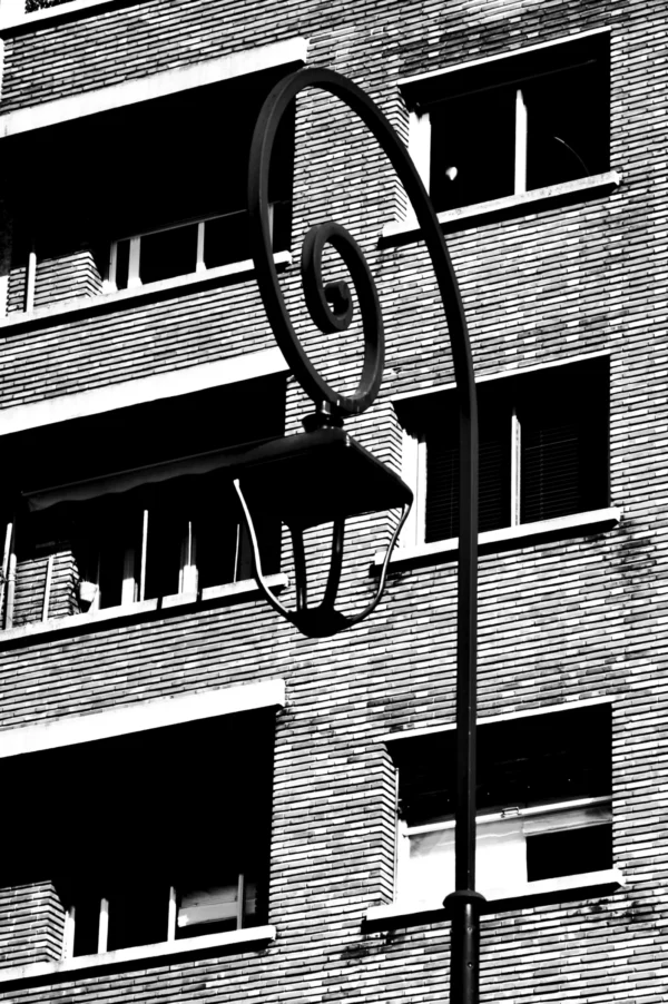 Thomas · 21/02/2026 · 2048 x 3080 Cette photographie met en scène un **réverbère vintage** à spirale, sujet principal. La composition est épurée, centrée sur l'objet avec un cadrage serré. L'arrière-plan montre une façade d'immeuble en briques, avec des fenêtres alignées.La lumière, contrastée, crée des ombres marquées, typique d'un cliché en **noir et blanc**. Les tons dominants varient du blanc au gris foncé, renforçant l'aspect dramatique. Le style, **minimaliste et graphique**, évoque une ambiance **urbaine et rétro**, presque mélancolique.