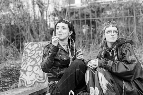 Laurence · 1920 x 1280 Cette photographie en noir et blanc met en scène deux jeunes femmes assises, semblant discuter ou se reposer. Le sujet principal est leur interaction, renforcée par leur proximité et leurs regards.La composition est naturelle, avec un cadrage serré sur elles, intégrant un fond flou de végétation et de graffiti. La lumière douce, probablement naturelle, crée des contrastes doux et des ombres marquées, accentuant les textures des vêtements et des visages.L’absence de couleurs et l’esthétique brute renforcent une ambiance intime, presque mélancolique, évoquant une scène de vie quotidienne authentique et spontanée.