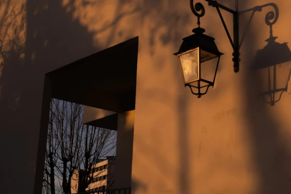 Laurence · 1920 x 1280 Cette photographie met en scène un **portique éclairé** avec deux lanternes anciennes comme sujet principal. La **composition** joue sur les contrastes entre ombres et lumières, avec un cadrage serré qui isole les éléments architecturaux.La **lumière dorée**, probablement celle du coucher de soleil, traverse l’encadrement, créant une ambiance chaleureuse et mystérieuse. Les **couleurs dominantes** sont des tons chauds (oranges, bruns), rehaussés par les reflets des lanternes.Le style est **minimaliste et poétique**, évoquant une atmosphère **nostalgique et sereine**, presque onirique.