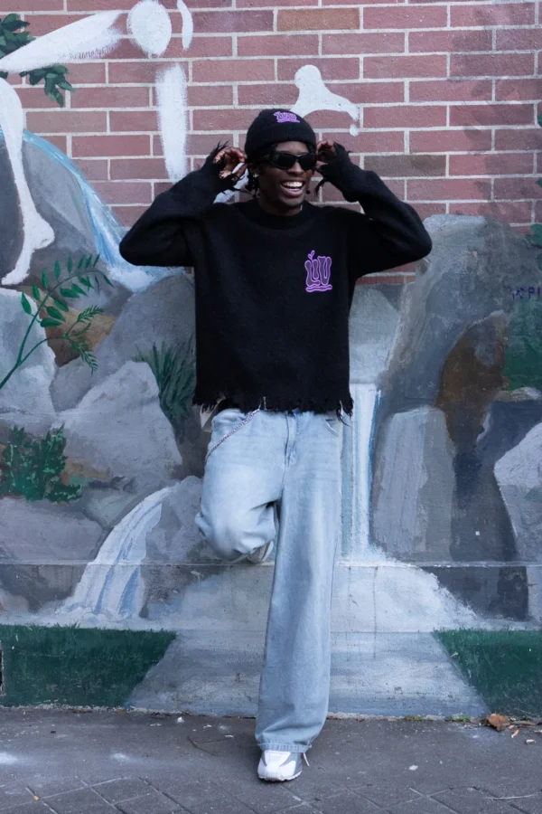 Laurence · 1280 x 1920 Cette photographie met en scène un jeune homme comme sujet principal, vêtu d'un sweat à capuche noir et de larges pantalons gris, posant devant un mur en brique et des graffitis.La composition est dynamique, avec un cadrage serré sur le sujet. La lumière semble naturelle, légèrement tamisée, créant des ombres douces. Les couleurs dominantes sont les tons froids (gris, noir) contrastant avec les touches de blanc et bleu des graffitis.Le style est urbain et décontracté, évoquant une ambiance streetwear. L'atmosphère est à la fois détendue et légèrement mystérieuse.
