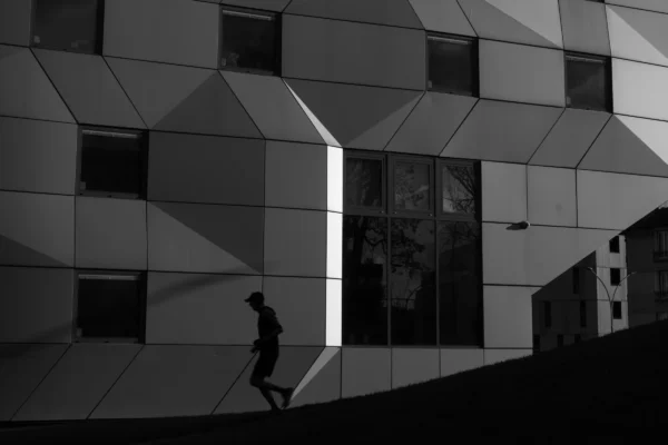 Don · 5324 x 3549 Cette photographie en noir et blanc met en scène une personne en mouvement devant un bâtiment moderne. Le sujet principal est le silhouetté, dynamisant la composition par son flou de mouvement.Le cadrage est serré, centré sur la personne et les lignes géométriques du bâtiment. La lumière, contrastée, accentue les ombres et les formes architecturales, créant une ambiance dramatique. Les tons dominants, du noir au gris, renforcent la modernité et la sobriété.Le style épuré et minimaliste évoque une atmosphère urbaine et contemplative, mêlant mouvement et immobilité.