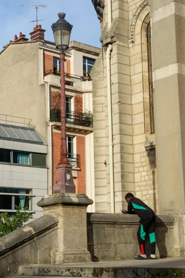 Cédric · 1365 x 2048 Cette photographie met en scène une personne, sujet principal, penchée sur une balustrade en pierre, vêtue d’un manteau vert et d’un pantalon coloré. La composition joue sur la verticalité avec un immeuble à balcons en arrière-plan et une église imposante.Le cadrage serré isole le personnage tout en intégrant des éléments architecturaux. La lumière naturelle, douce et légèrement contrastée, éclaire la scène sans ombre marquée. Les couleurs dominantes sont les tons neutres (gris, beige) adoucis par les touches de vert et de rouge des balcons.Le style est documentaire et réaliste, créant une ambiance urbaine paisible et légèrement mélancolique.