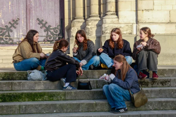 Cédric · 2048 x 1365 Cette photographie montre un groupe de cinq jeunes femmes assises sur des marches d’escalier extérieur. Le sujet principal est leur interaction conviviale et détendue.La composition est dynamique, avec un cadrage serré qui met en valeur les visages et les gestes. La lumière naturelle, douce et légèrement tamisée, crée des ombres douces. Les couleurs dominantes sont les tons neutres (beige, gris) et les couleurs naturelles (marron, bleu), renforçant une ambiance chaleureuse et informelle.Le style est documentaire, capturant un moment authentique et spontané, évoquant une ambiance amicale et décontractée.