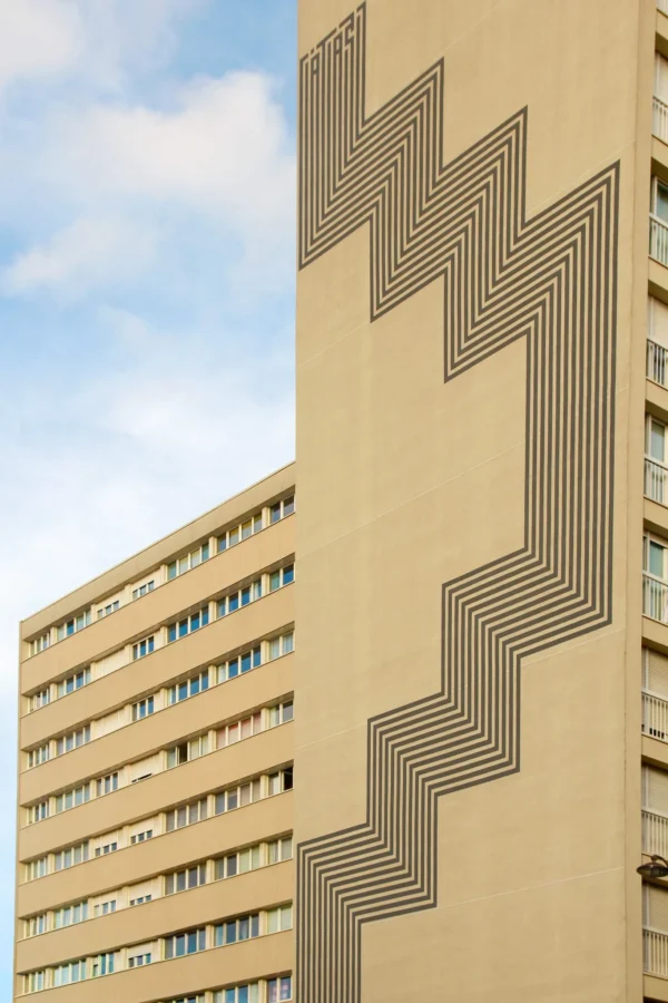Cédric · 1365 x 2048 Cette photographie met en scène deux bâtiments modernes. Le sujet principal est le bâtiment de droite, marqué par une façade géométrique en forme de zigzag métallique. La composition joue sur le contraste entre les lignes épurées et les formes architecturales. Le cadrage est serré, mettant en valeur les détails architecturaux.La lumière est naturelle, éclairant uniformément la scène. Les couleurs dominantes sont des tons beiges et gris, avec des touches métalliques argentées. Le style est architectural et minimaliste, évoquant une ambiance urbaine et contemporaine, presque froide mais élégante.