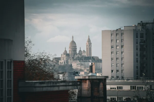 Cédric · 2048 x 1365 Cette photographie met en scène une ville avec, en arrière-plan, une cathédrale gothique emblématique, probablement Notre-Dame de Paris, partiellement visible à travers les bâtiments modernes.La composition joue sur le contraste entre l'architecture ancienne et contemporaine. Le cadrage est serré, avec des bâtiments en premier plan qui encadrent la vue. La lumière est douce et tamisée, créant une ambiance mélancolique et nostalgique. Les couleurs dominantes sont des tons gris et beiges, adoucis par des touches de vert et de bleu dans le ciel.Le style est réaliste et légèrement flou, renforçant une atmosphère contemplative et poétique.