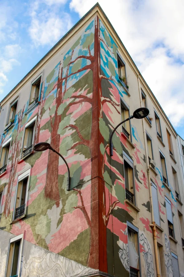 Cédric · 1365 x 2048 Cette photographie met en scène un immeuble dont la façade est recouverte d’un **mural végétal stylisé**. Le **sujet principal** est cette fresque colorée, représentant des branches et des feuilles aux teintes vives.La **composition** est dynamique, avec un **cadrage en contre-plongée** accentuant la hauteur du bâtiment. La **lumière** naturelle, probablement ensoleillée, éclaire les couleurs et crée des contrastes nets. Les **couleurs dominantes** sont des verts, roses et ocres, apportant une touche vibrante et organique.Le **style** est artistique et urbain, évoquant un mélange de nature et d’architecture. L’**ambiance** est à la fois moderne et poétique, dynamisant le paysage urbain.