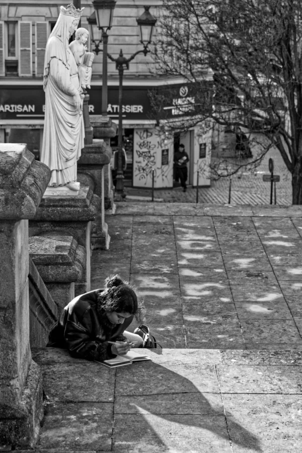 Mickaël · 07/02/2026 · 2907 x 4355 Cette photographie en noir et blanc met en scène une femme assise sur des marches, penchée en avant, semblant écrire ou dessiner. Le sujet principal est cette personne, isolée dans un espace urbain.La composition repose sur une perspective descendante, avec des escaliers menant vers elle. À l’arrière-plan, des statues et des enseignes ajoutent une touche historique. La lumière est douce, créant des contrastes subtils. Le style est documentaire, capturant un moment intime dans un cadre public. L’ambiance est à la fois mélancolique et contemplative, renforcée par le monochrome.