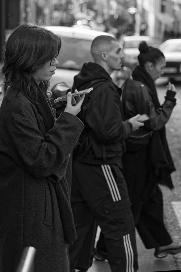 Mickaël · 07/02/2026 · 2633 x 3944 Cette photographie en noir et blanc montre trois personnes en extérieur, dont deux hommes et une femme en premier plan. Le sujet principal semble être la femme de profil, absorbée par son téléphone.La composition est dynamique, avec un cadrage serré sur les visages et les mains. La lumière est naturelle, douce mais contrastée, créant des ombres marquées. L’ambiance est urbaine et moderne, reflétant une vie quotidienne intense. Le style est documentaire, capturant un instant réel et spontané. Les couleurs, absentes en raison du noir et blanc, laissent place à des jeux de tons clairs et sombres.