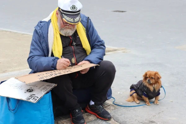 Vera · 6960 x 4640 Cette photographie met en scène un homme assis, vêtu d’un uniforme marin (veste bleue, béret et écharpe jaune), occupé à écrire sur une feuille posée sur ses genoux. À ses côtés, un petit chien est attaché par une laisse.La composition est simple et naturelle, avec un cadrage serré sur les deux sujets. La lumière semble douce et naturelle, probablement en extérieur. Les couleurs dominantes sont les tons froids (bleu de la veste, gris du sol) et chauds (jaune de l’écharpe, brun du chien). L’ambiance est calme et quotidienne, évoquant une scène de vie paisible, peut-être portuaire.
