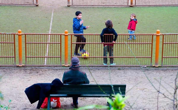 Pascale · 1000 x 617 Cette photographie capture une scène urbaine au parc. Le sujet principal est un homme assis sur un banc, tourné vers la gauche, avec un sac à ses pieds. En arrière-plan, trois enfants jouent près d'une clôture jaune.La composition est équilibrée, avec une ligne horizontale marquée par le banc et la clôture. Le cadrage est large, incluant un espace vert et une partie du ciel. La lumière semble naturelle, mais légèrement voilée, créant une ambiance douce et un peu grise.Les couleurs dominantes sont les tons froids (gris, bleu) avec des touches de jaune vif (clôture) et de vert (végétation). Le style est réaliste et documentaire, évoquant une ambiance calme et quotidienne.