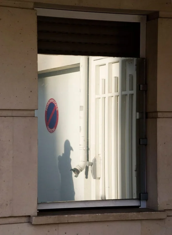 Pascale · 733 x 1000 Cette photographie montre une silhouette floue à travers une fenêtre ouverte. Le sujet principal est cette ombre indistincte, probablement une personne, en contre-jour.La composition est simple et épurée, avec un cadrage serré sur la fenêtre. La lumière, vive et naturelle, provient de l’extérieur, créant des contrastes marqués et des reflets sur la vitre. Les couleurs dominantes sont les tons clairs (blanc, gris) et une touche de rouge due au panneau circulaire partiellement visible.Le style est minimaliste et documentaire, évoquant une ambiance mystérieuse et légèrement mélancolique.