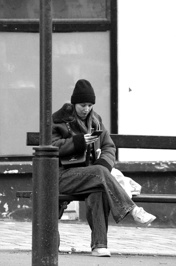 Pascale · 664 x 1000 Cette photographie en noir et blanc montre un homme assis sur un banc, sujet principal. Il semble absorbé par son téléphone portable, vêtu d’un bonnet et d’une veste, évoquant un style urbain et décontracté.La composition est simple et équilibrée, avec un cadrage serré sur le sujet, encadré par un poteau et une vitre en arrière-plan. La lumière est douce, probablement naturelle, créant des contrastes modérés. L’ambiance est calme et introspective, renforcée par le monochrome qui accentue les textures et les formes.