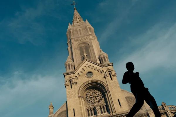 Don · 6706 x 4471 Cette photographie met en scène un **silhouette humaine** en mouvement devant un **édifice religieux** imposant, probablement une cathédrale gothique. La **composition** joue sur le contraste entre la silhouette sombre et l’architecture claire.Le **cadrage** est serré sur la personne, avec la cathédrale en arrière-plan. La **lumière** est douce, créant un effet dramatique avec des ombres marquées. Les **couleurs dominantes** sont les tons beiges et blancs de la cathédrale, contrastant avec le bleu du ciel.Le style est **cinématographique**, évoquant une ambiance **mystérieuse et poétique**, renforcée par les jeux d’ombres et de lumière.