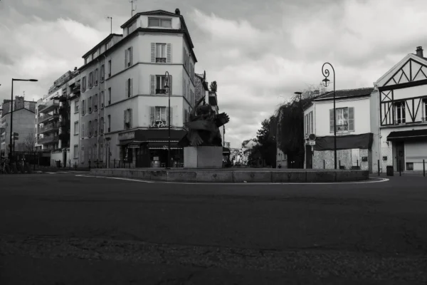 Don · 7008 x 4672 Cette photographie en noir et blanc capture une intersection urbaine déserte. Le sujet principal est un immeuble à l’architecture classique, partiellement masqué par une structure en construction. La composition met en valeur les lignes architecturales et les perspectives des bâtiments environnants.Le cadrage est large, incluant une rue vide et des bâtiments secondaires. La lumière, tamisée, crée des contrastes doux entre ombres et clartés, renforçant l’ambiance mélancolique et calme. Les tons dominants, en niveaux de gris, accentuent la texture des matériaux et l’atmosphère nostalgique. Le style est documentaire, évoquant une ville en transition.