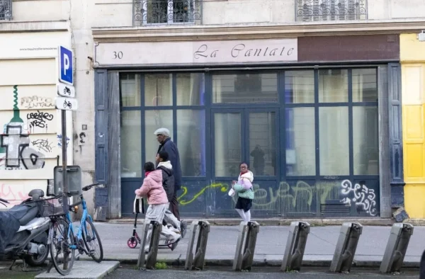 Muriel · 1080 x 709 Cette photographie montre une scène urbaine devant une boutique nommée "Le Santet". Le sujet principal est une femme poussant un enfant en trottinette, accompagnée d’un autre enfant courant vers eux.La composition est dynamique, avec un cadrage serré sur le trottoir et les personnes. Les couleurs dominantes sont neutres (gris, beige) avec des touches de bleu et de vert. La lumière est naturelle, légèrement tamisée, créant une ambiance calme mais vivante.Le style est documentaire, reflétant un instant de vie quotidienne en ville. L’ambiance est celle d’un quartier résidentiel animé.
