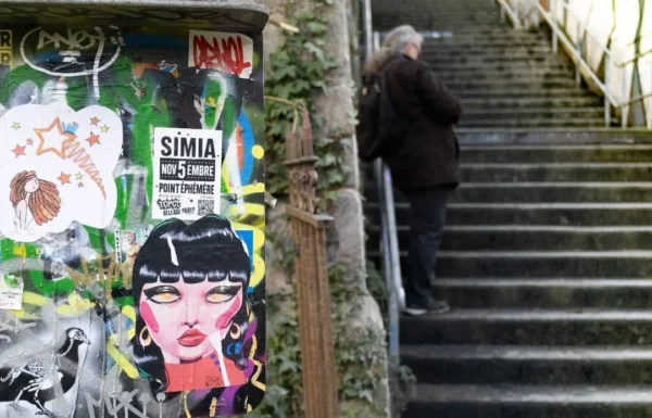 Muriel · 1080 x 693 Cette photographie montre un **mur tagué** avec des affiches colorées, dont une figure féminine stylisée et un texte partiellement lisible ("SIMIA"). En arrière-plan, une personne descend un escalier en pierre.La **composition** oppose l’art urbain dynamique au calme des marches. Le **cadrage** est serré sur le mur, avec une ouverture sur l’escalier. La **lumière** est naturelle, douce mais contrastée, créant des ombres marquées. Les **couleurs dominantes** sont vives (rouge, bleu, noir) sur le mur, contrastant avec les tons neutres des pierres.Le **style** est urbain et artistique, évoquant une **ambiance** à la fois vivante et mélancolique, entre street art et solitude.