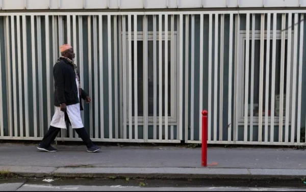 Muriel · 1080 x 678 Cette photographie met en scène un homme en mouvement, vêtu d’un manteau sombre, portant des documents. Le sujet principal est flou, suggérant un cadrage dynamique.La composition repose sur une perspective basse, avec un premier plan occupé par un poteau rouge vif. Les barrières métalliques en arrière-plan structurent l’espace. La lumière est naturelle, mais tamisée, créant des contrastes doux.Les couleurs dominantes sont les tons gris et verts métalliques, adoucis par le manteau noir de l’homme. Une touche de rouge vif (le poteau) apporte un contraste saisissant.Le style est sobre et réaliste, évoquant une ambiance urbaine et contemporaine, presque mélancolique.