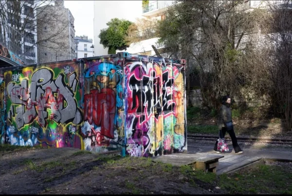 Muriel · 1080 x 727 Cette photographie met en scène un **mur tagué** couvert de graffitis colorés et variés, formant le sujet principal. La composition montre une **perspective légèrement basse**, encadrée par des arbres et des bâtiments en arrière-plan.La lumière est naturelle, douce, avec des ombres discrètes, suggérant un jour nuageux. Les **couleurs dominantes** sont vives et contrastées : rouge, bleu, violet, jaune et vert, typiques des graffitis urbains.Le style est **urbain et artistique**, reflétant une culture street art. L’ambiance est à la fois **dynamique et mélancolique**, mêlant énergie créative et solitude, renforcée par la présence d’une personne assise, isolée.