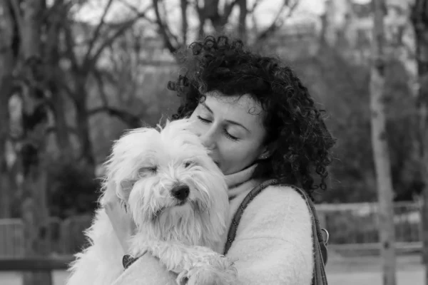 Vera · 5946 x 3964 Cette photographie met en scène une femme comme sujet principal, serrant contre elle un petit chien à poils longs. La composition est centrée et intime, avec un cadrage serré sur les deux sujets. La lumière est douce et naturelle, probablement en extérieur, créant des contrastes doux.En noir et blanc, les tons dominants varient du clair au foncé, mettant en valeur les textures des cheveux de la femme et du pelage du chien. Le style est simple et émotionnel, évoquant une ambiance chaleureuse et affectueuse, renforcée par le contact physique entre la femme et son animal.