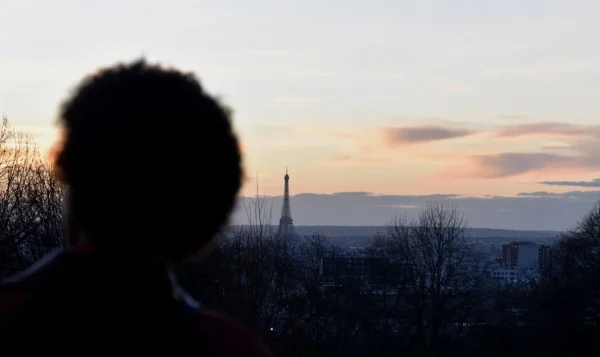 Franck T. · 1400 x 833 Cette photographie présente un **sujet principal flou** vu de dos, dont seul le contour de la tête est visible. Le **cadrage** met en valeur un **paysage urbain** avec, au loin, la silhouette emblématique de la **tour Eiffel**. La **composition** joue sur le contraste entre le premier plan sombre et l’arrière-plan lumineux.La **lumière**, douce et dorée, évoque un **crépuscule**, tandis que les **couleurs dominantes** sont des teintes chaudes (oranges, roses) et des tons froids (bleus grisâtres). Le style est **minimaliste et poétique**, créant une **ambiance mélancolique et contemplative**.