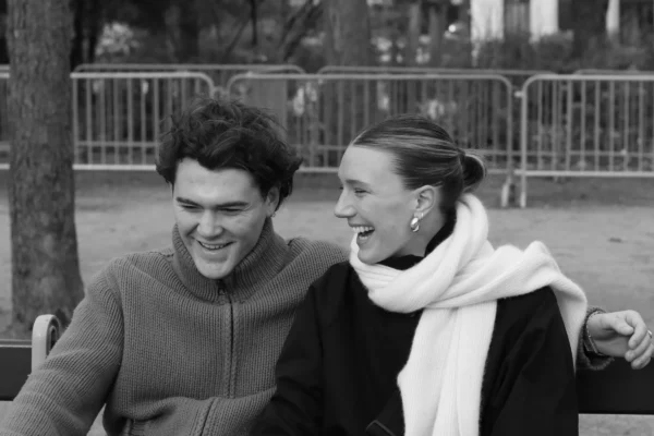 Vera · 6030 x 4020 Cette photographie en noir et blanc met en scène deux jeunes gens comme sujets principaux, souriants et complices. La composition est naturelle, avec un cadrage serré qui les place au centre. L’arrière-plan flou montre une barrière et un espace vert, suggérant un parc.La lumière est douce, probablement naturelle, créant des contrastes modérés. Les couleurs dominantes, en niveaux de gris, renforcent l’aspect intemporel et élégant. Le style est spontané et chaleureux, évoquant une ambiance joyeuse et détendue, presque nostalgique.