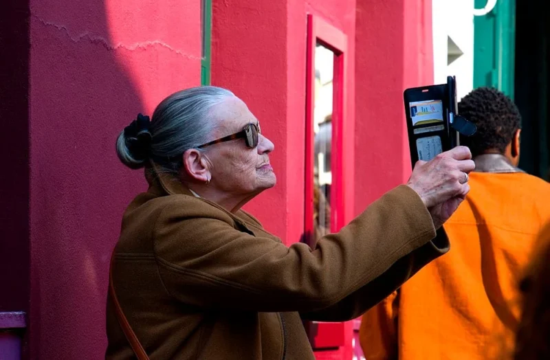 Pascale · 21/03/2026 · 1000 x 655 Cette photographie met en scène une personne âgée, sujet principal, prenant une photo d’un homme en orange avec un appareil rétro. La composition est serrée, centrée sur les deux individus. Le cadrage est en plan serré, capturant leurs interactions.La lumière est naturelle et semble provenir d’une source latérale, créant des ombres douces. Les couleurs dominantes sont le rouge vif du mur, l’orange du vêtement de l’homme et les tons neutres du manteau de la personne âgée. Le style est documentaire et spontané, évoquant une ambiance authentique et urbaine.